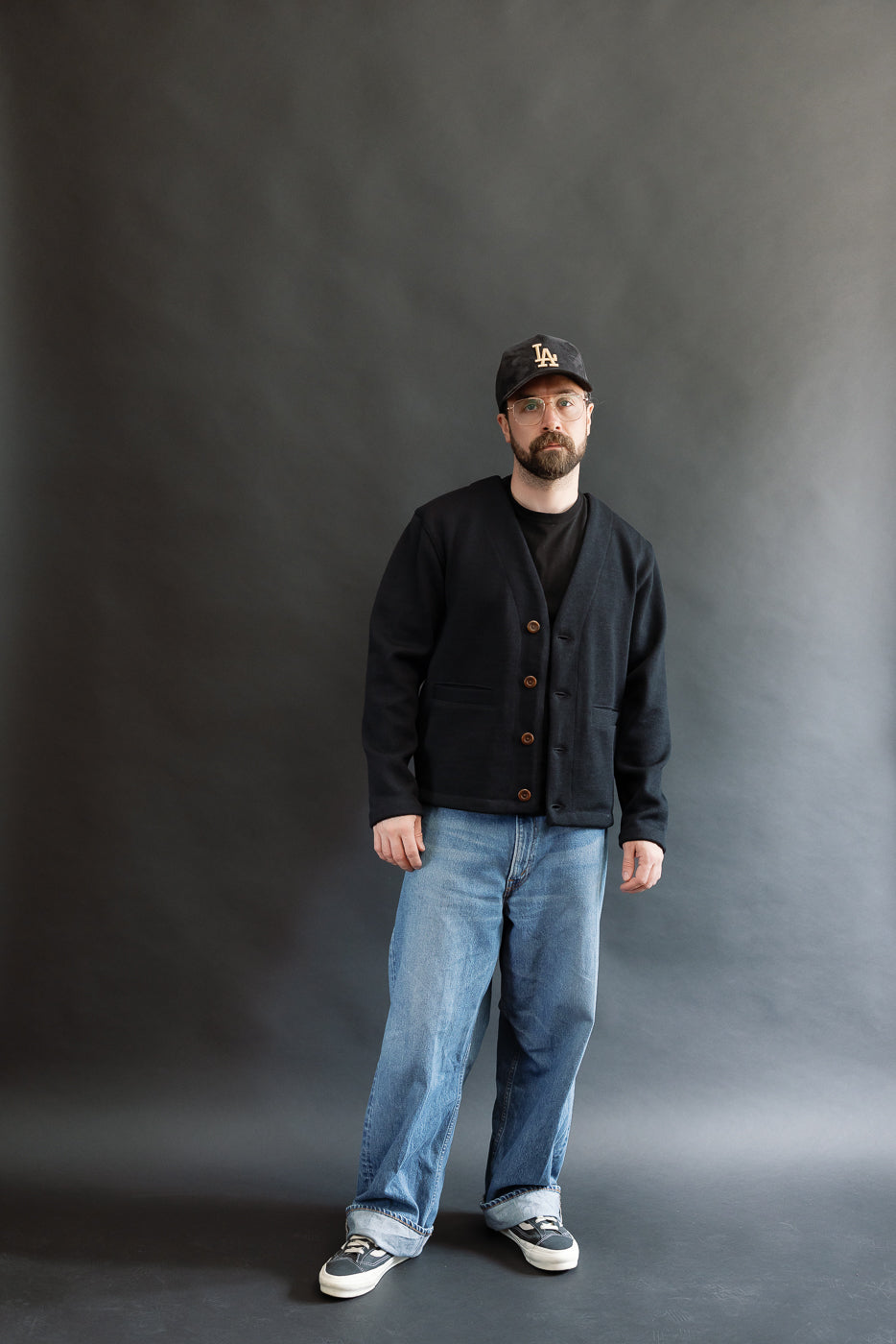 Male model wearing Black Dant slouchy cardigan with brown buttons and washed baggy blue jeans and black cap, full body