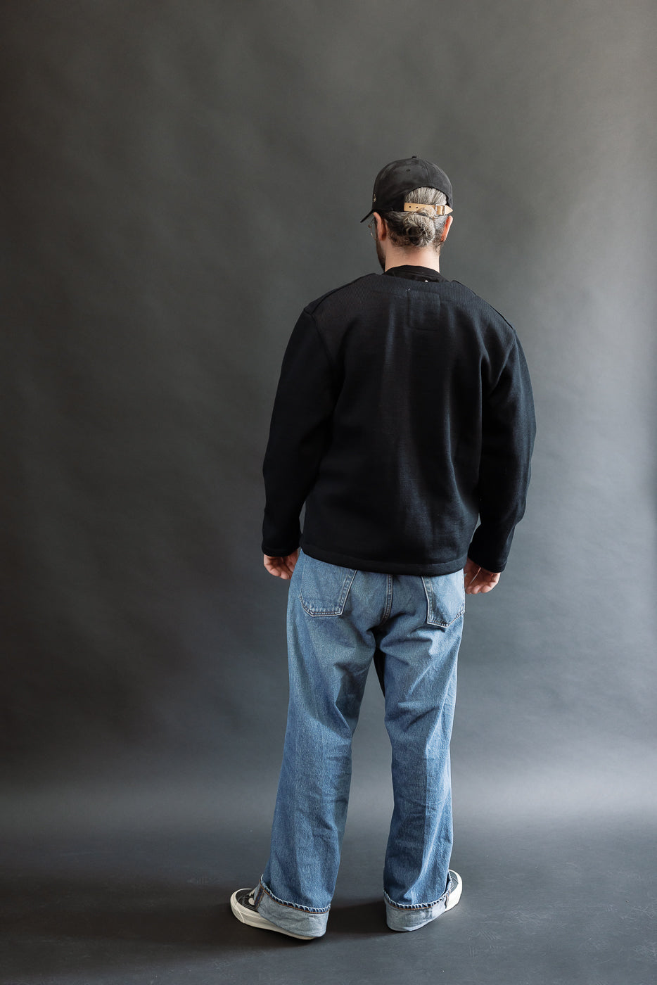 Male model wearing Black Dant slouchy cardigan with brown buttons and washed baggy blue jeans and black cap, full body, back view