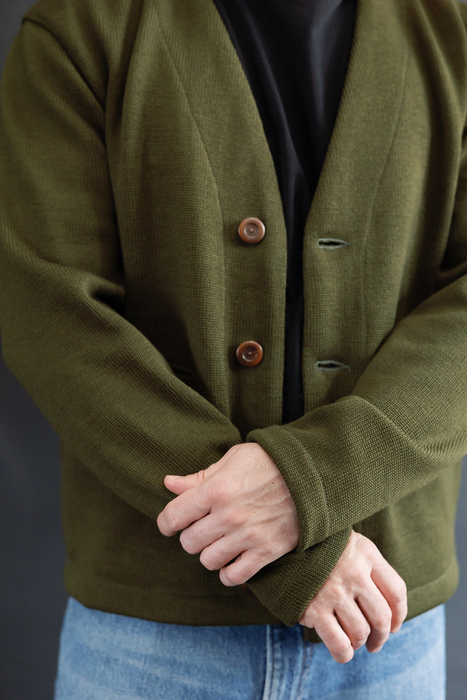close up of person wearing a black shirt with an olive green cardigan with brown corozo buttons on a dark background