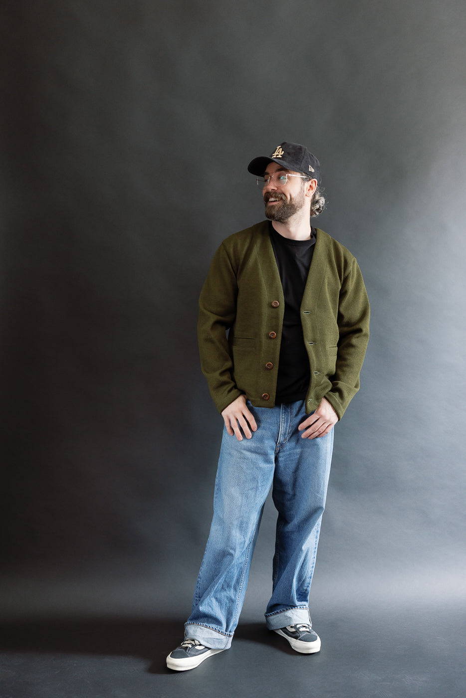 Male model wearing an olive green slouchy cardigan, black cap, and baggy washed blue jeans against a black background, full body, front view
