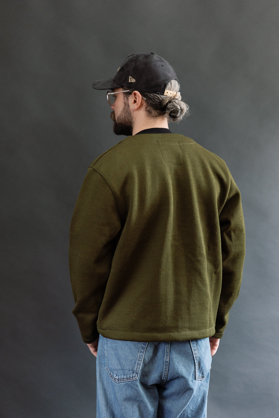 Male model wearing an olive green slouchy cardigan w black t-shirt, black cap, 3/4 length, back view