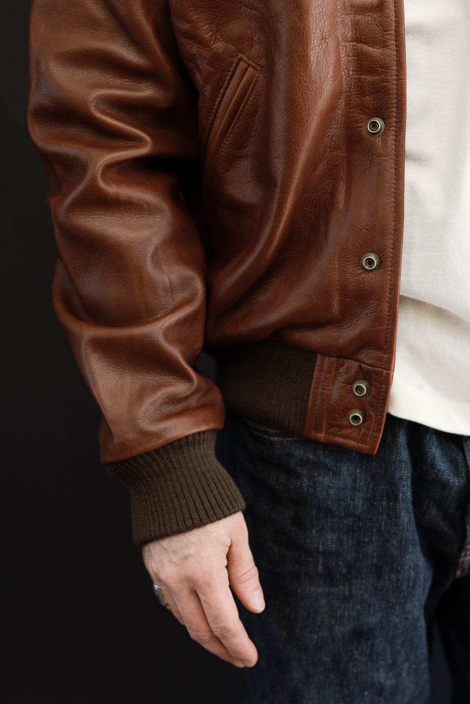 Close-up side view of a brown leather bomber jacket with ribbed cuffs and hem on a male model.