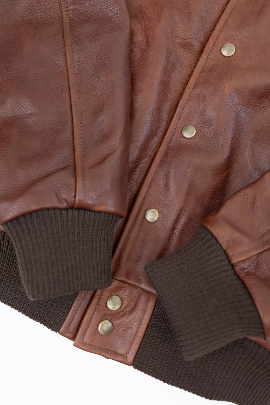 Close-up front view of brown leather jacket showing texture, snap buttons, and ribbed cuffs.