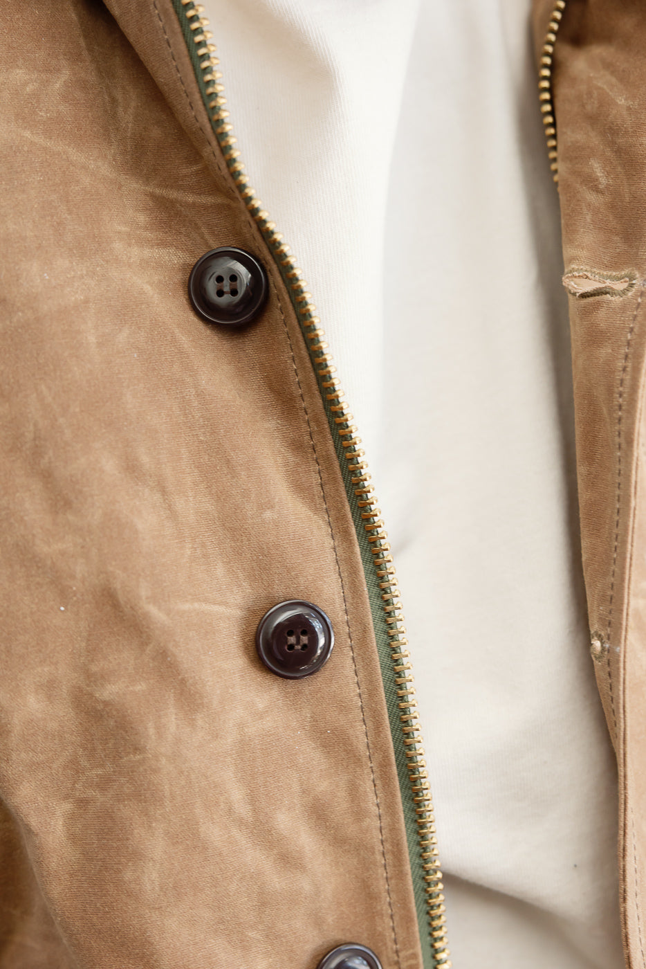 Close-up of the front right side of a brown waxed canvas jacket showing brass zipper and dark buttons.