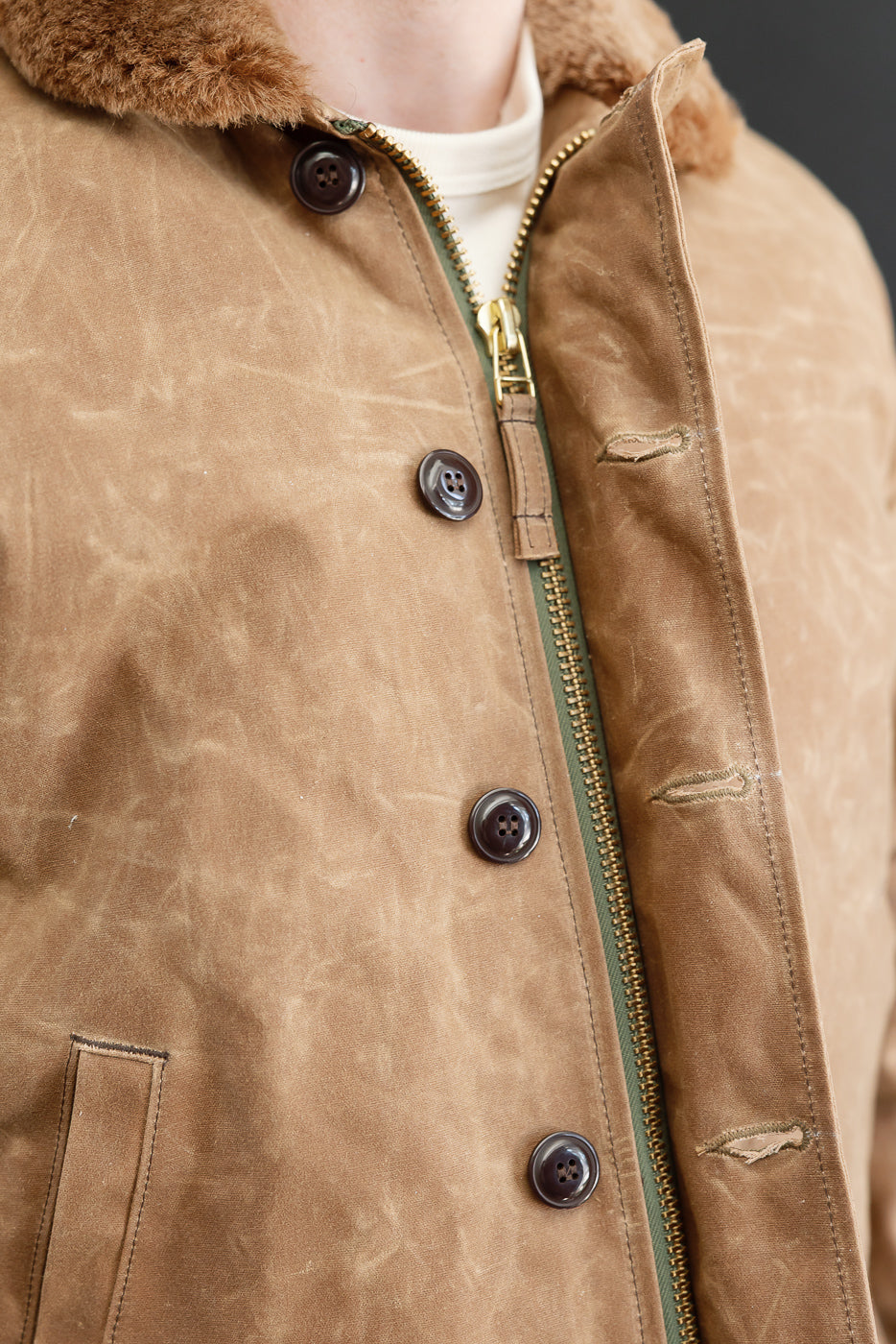 Close-up front view of a tan canvas jacket showing brass zipper, brown buttons, and faux fur collar.