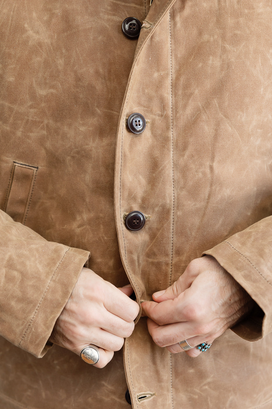 Close-up front view of a male model's hands fastening a brown waxed canvas jacket with dark buttons.