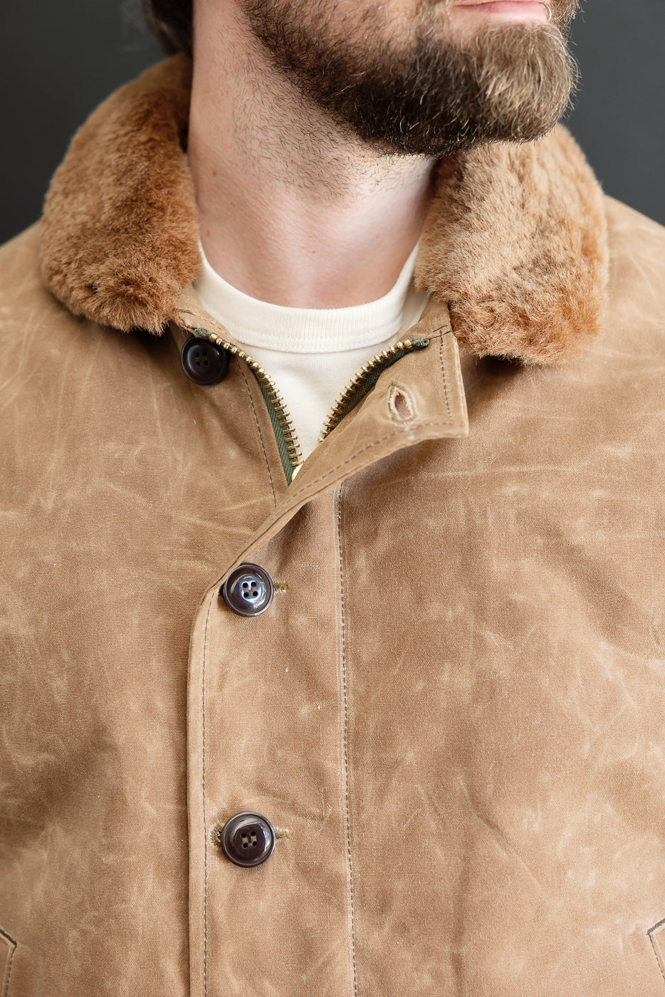 Close-up front view of a male model wearing a tan canvas jacket with a brown shearling collar and dark buttons.
