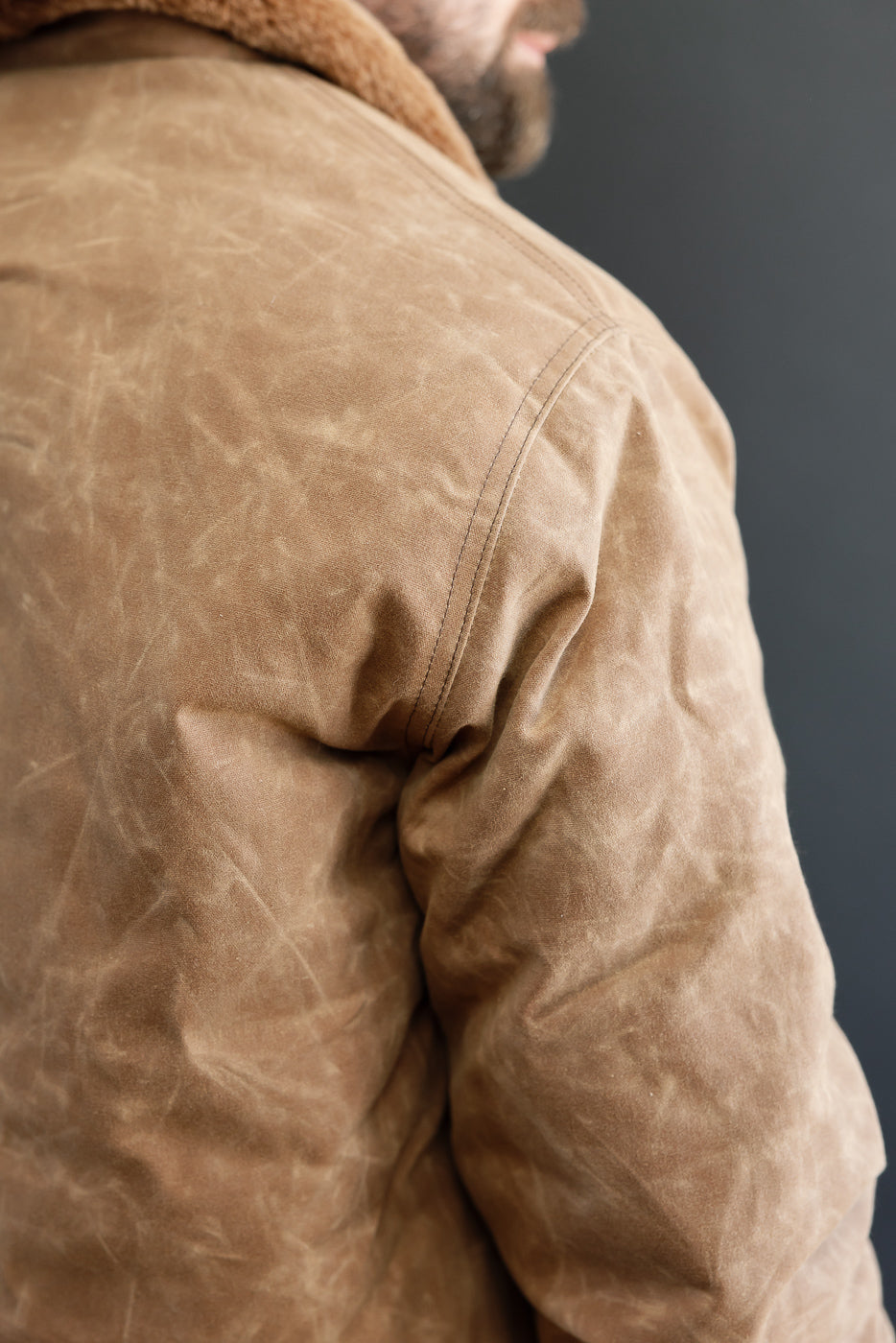 Close-up back view of a tan waxed canvas jacket showing fabric texture and shoulder seam.