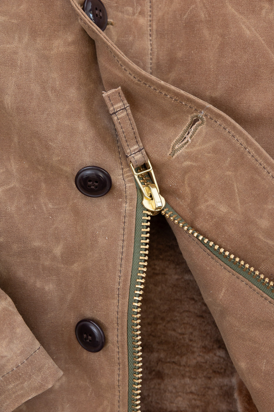 Close-up of the front center of a brown waxed canvas jacket showing a brass zipper and buttons.