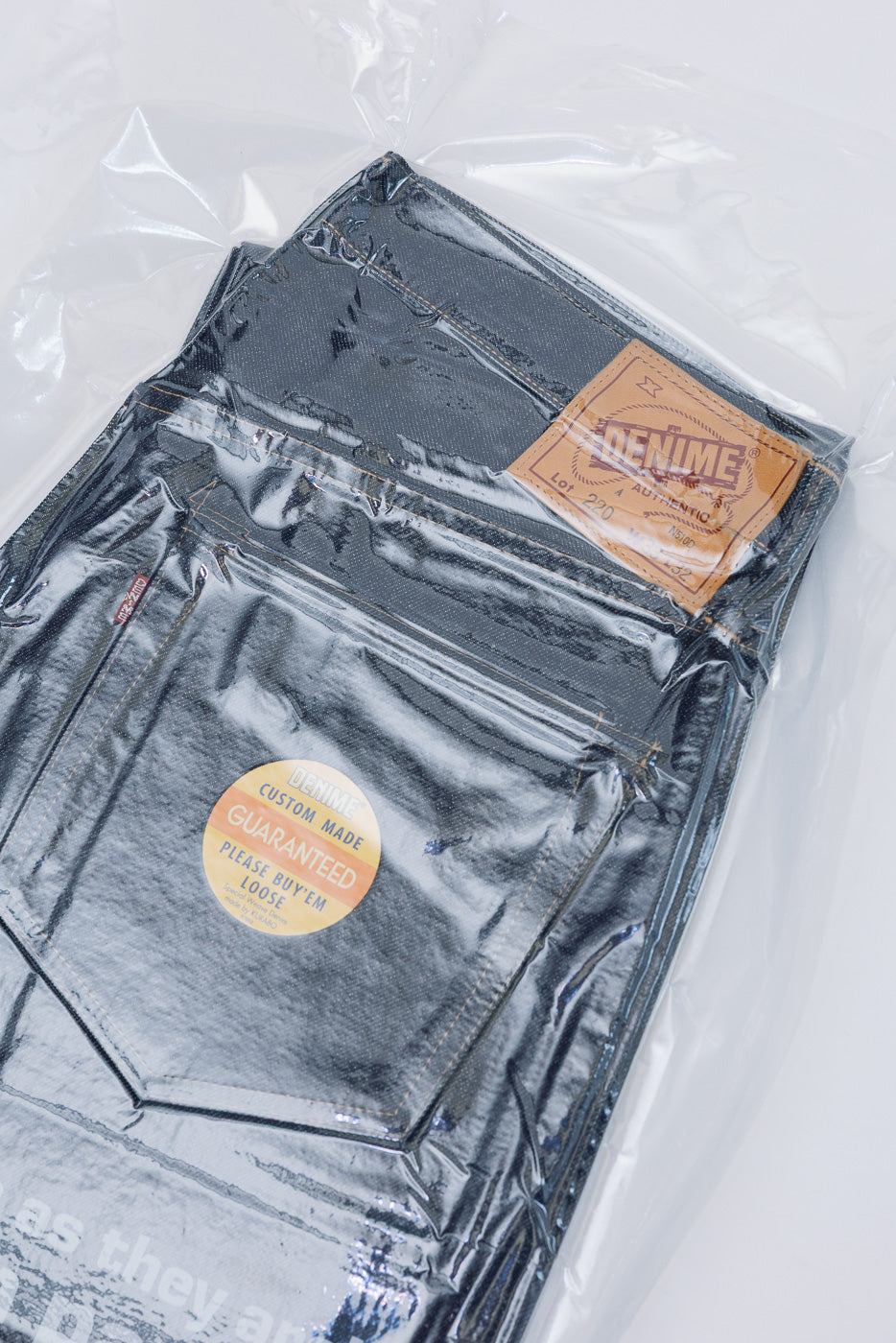 Flat lay, back view of dark selvedge denim jeans in plastic packaging with leather patch.