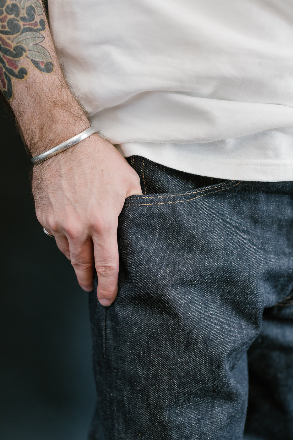 Person wearing a white shirt and dark jeans with a tattoo on the arm.
