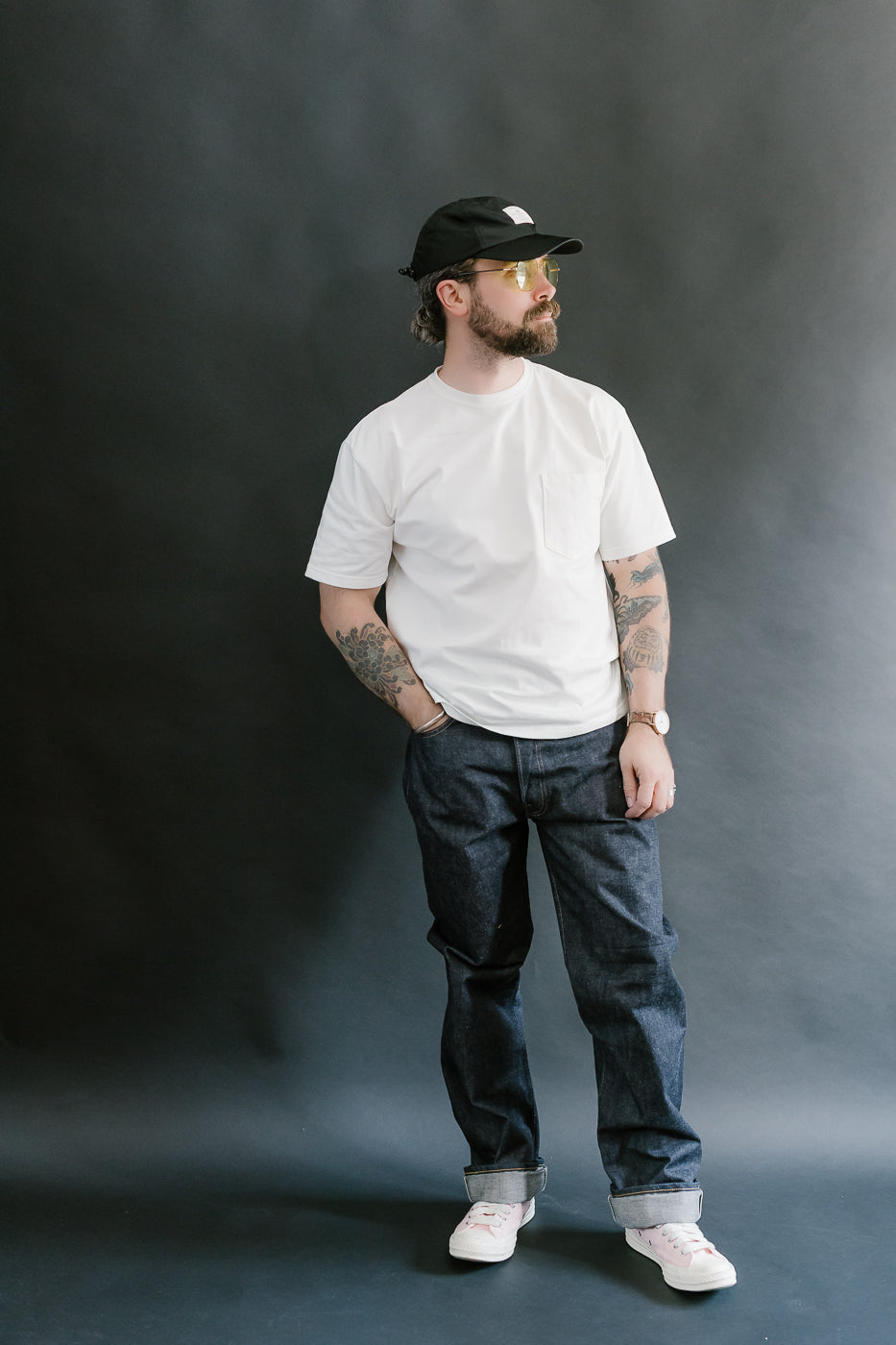 Man wearing a white t-shirt, dark jeans, and a black cap against a gray background