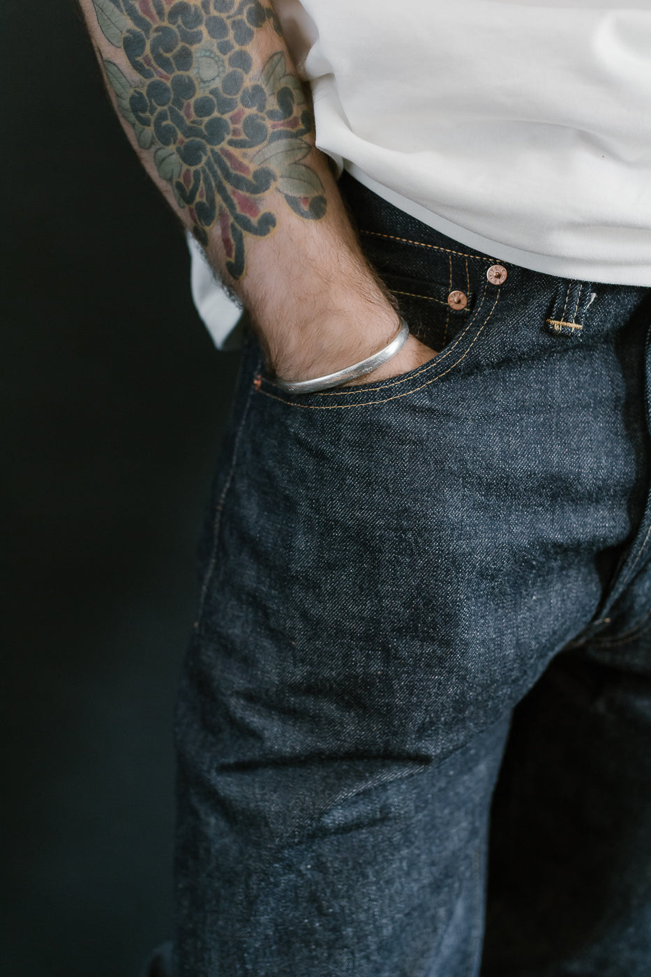 Person wearing blue jeans with a tattooed arm on a dark background