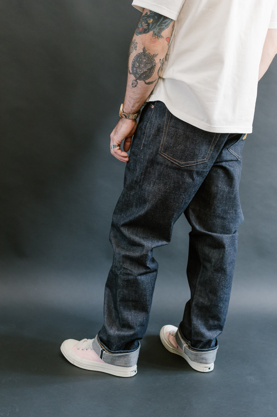 Person wearing dark jeans and a white shirt with tattoos on arms against a gray background