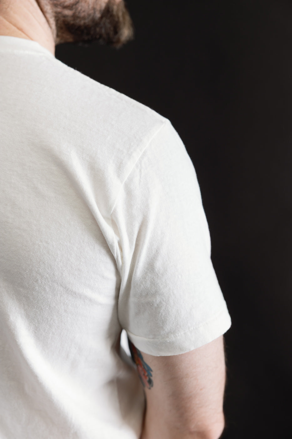 Back view of a 5.4oz Raffy Tenjiku Printed T-Shirt Grotius - Off White, showcasing its seamless tubular construction and slub texture against a dark backdrop, with a tattoo partially visible on the arm.