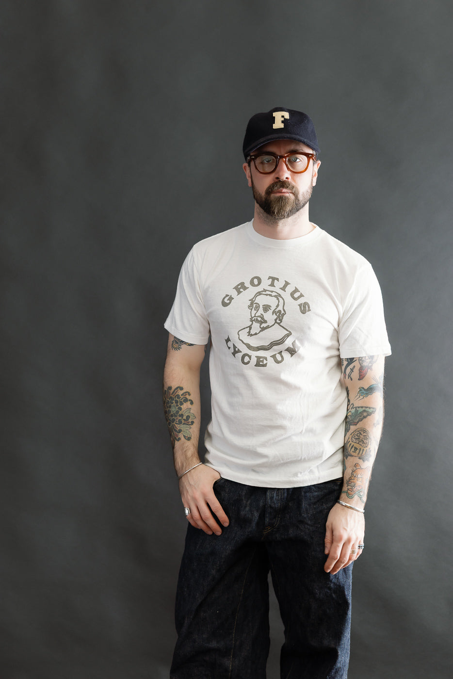 A casually dressed man models the 5.4oz Raffy Tenjiku Printed T-Shirt Grotius - Off White, featuring a stylized portrait and GROTIUS LYCEUM text, paired with dark blue jeans.