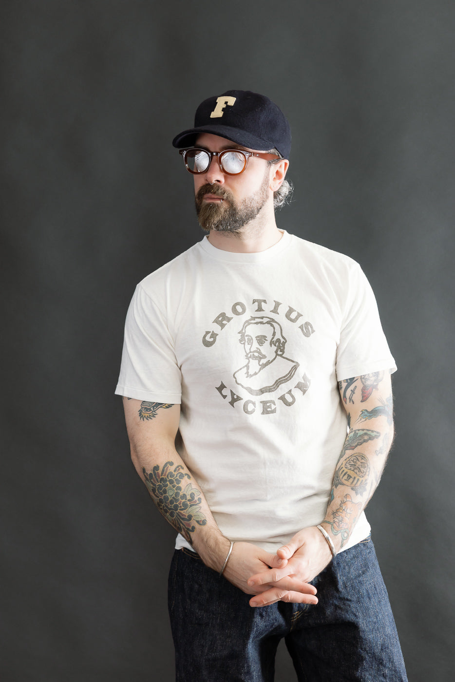 Man wearing 5.4oz - Raffy Tenjiku Printed T-Shirt Grotius - Off White, featuring GROTIUS LYCEUM text and portrait, styled with dark denim jeans, round eyeglasses, and a black baseball cap.
