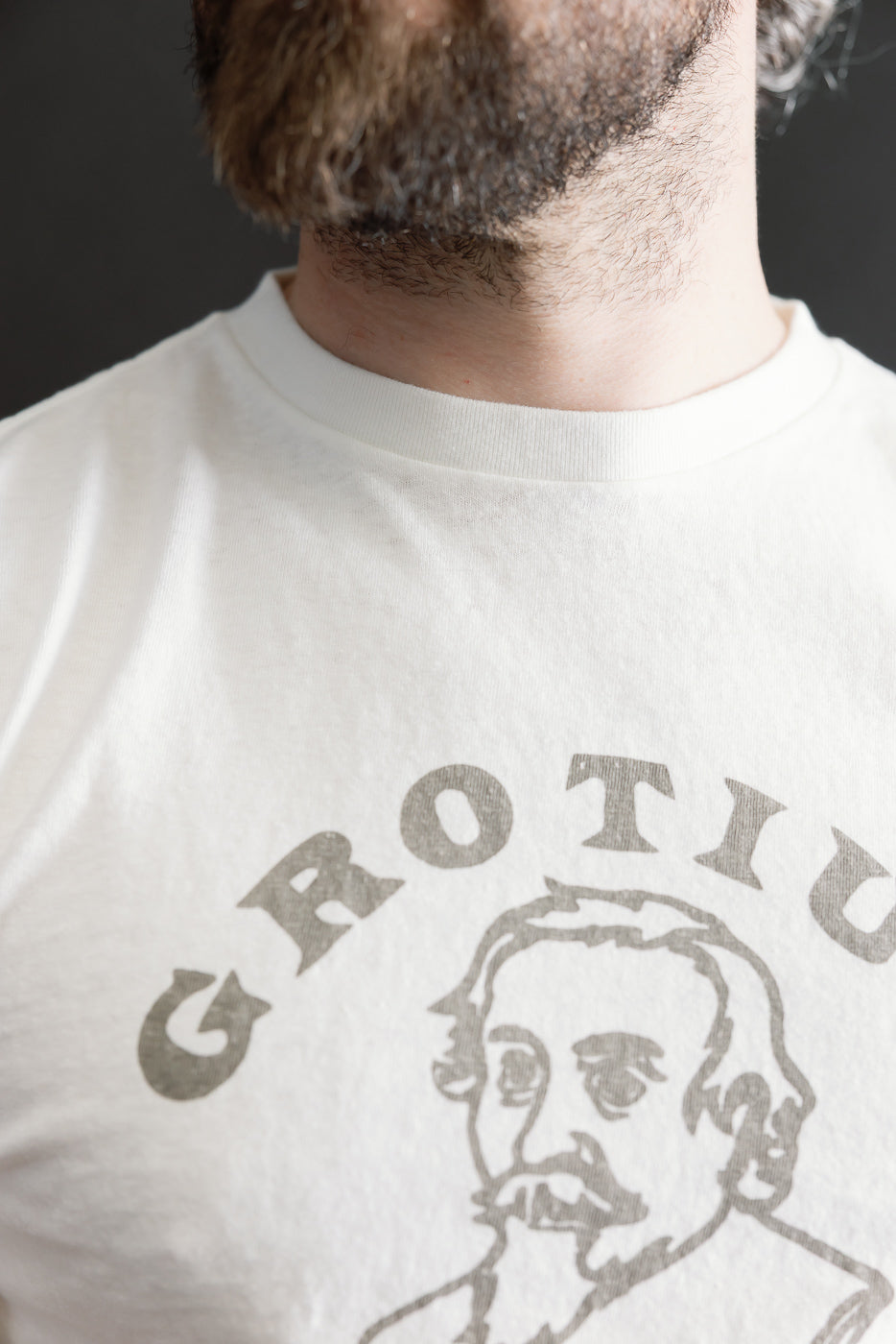Close-up of a man wearing the 5.4oz Raffy Tenjiku Printed T-Shirt Grotius - Off White, showcasing a stylized bearded portrait design and emphasizing the tubular knit fabric's unique nep-like texture.