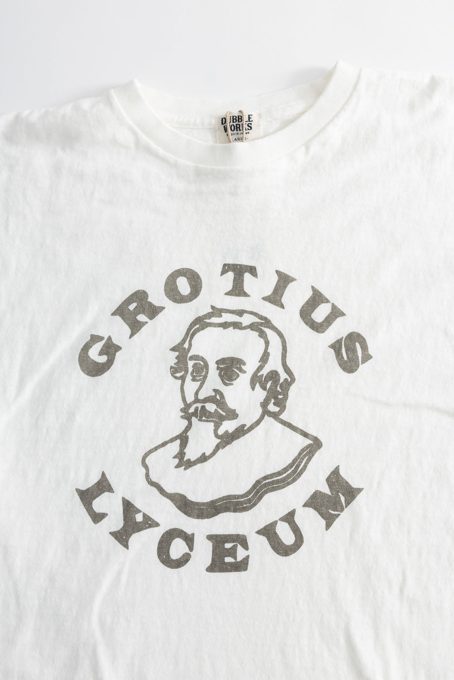 5.4oz Raffy Tenjiku Printed T-Shirt Grotius - Off White, featuring a monochrome line-drawing and serif text, laid flat to show its seam-free tubular knit and textured slub jersey fabric.