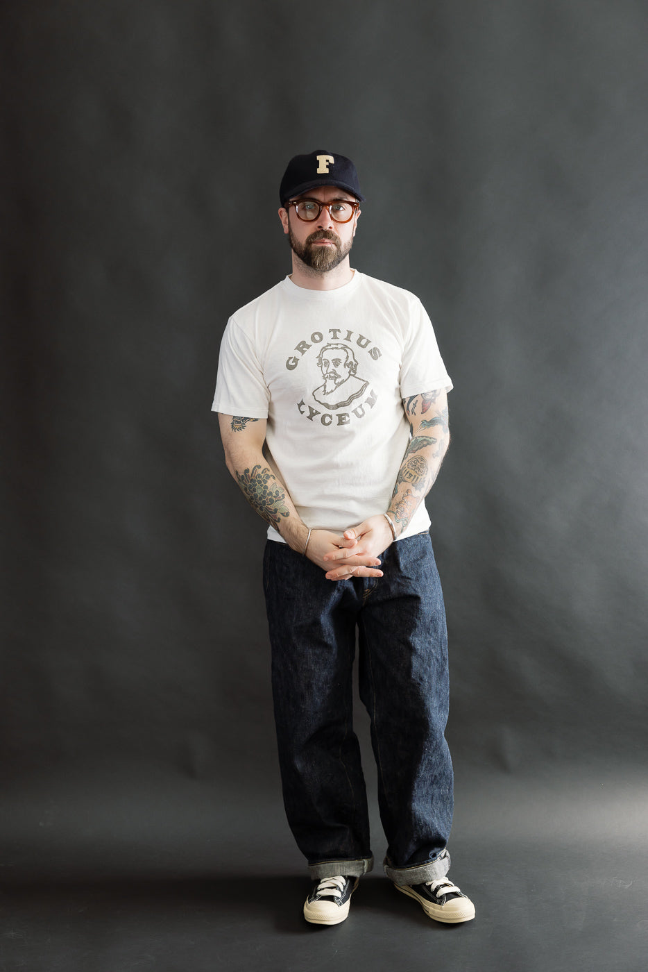 Man wearing 5.4oz Raffy Tenjiku Printed T-Shirt Grotius - Off White, paired with dark indigo jeans, black-and-white sneakers, and a cap, showcasing tattoos. The T-shirt features a vintage-inspired design.