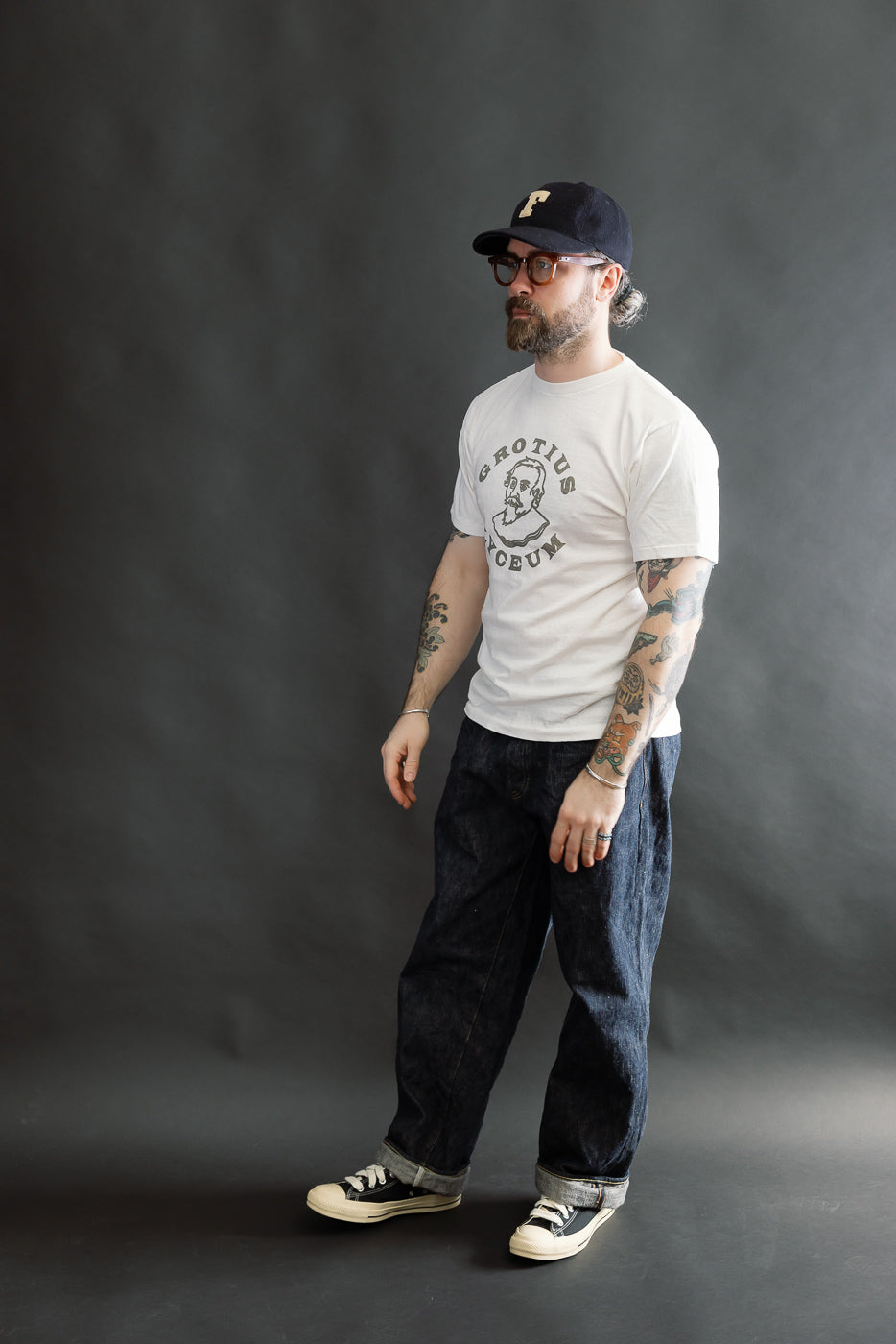 A bearded man in a dark cap models the 5.4oz Raffy Tenjiku Printed T-Shirt Grotius in off-white, showcasing its tubular knit and colorful forearm tattoos against a neutral backdrop.