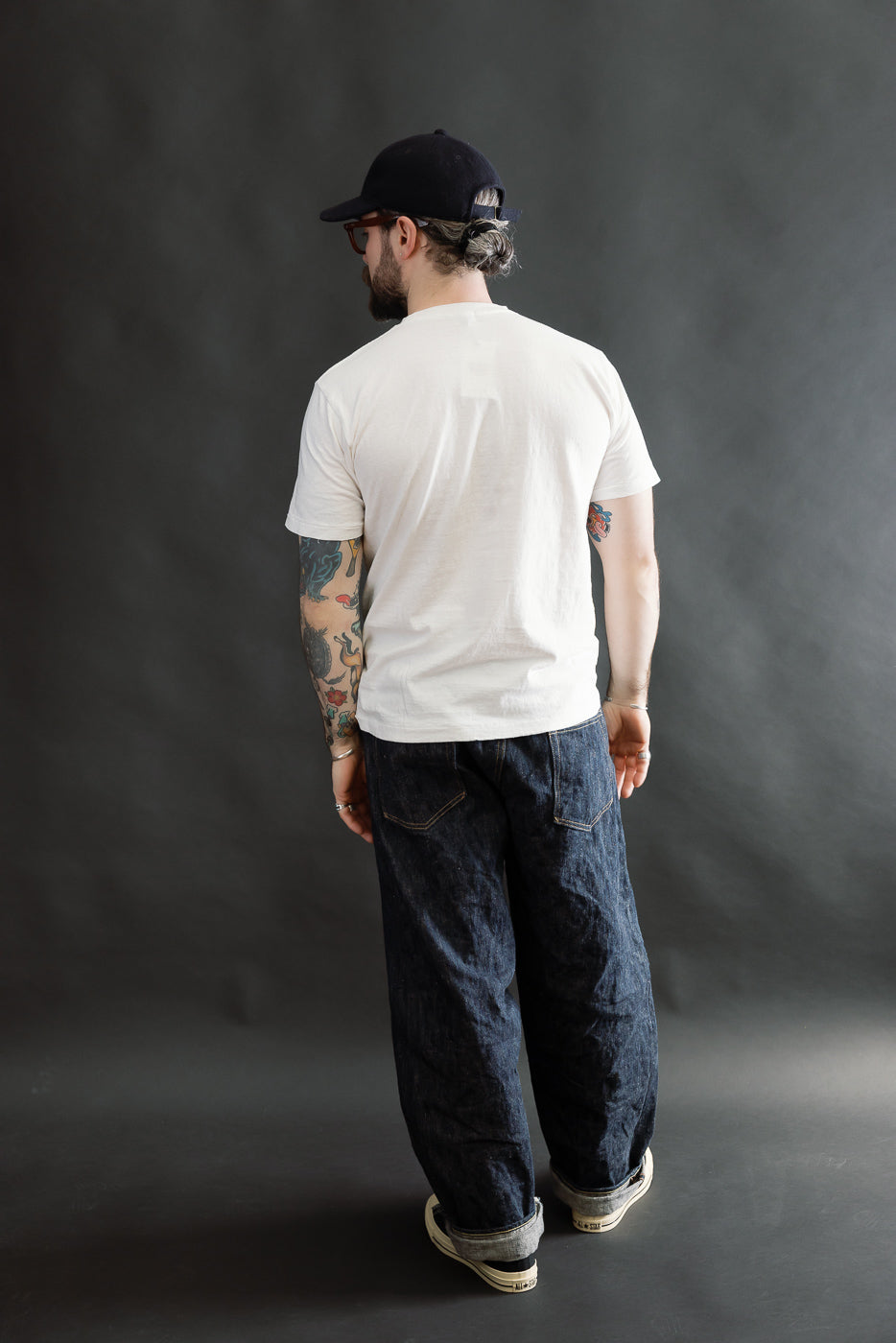 A man models the 5.4oz Raffy Tenjiku Printed T-Shirt Grotius - Off White, showcasing its seamless tubular knit and vintage-inspired design, paired with indigo denim and off-white sneakers.