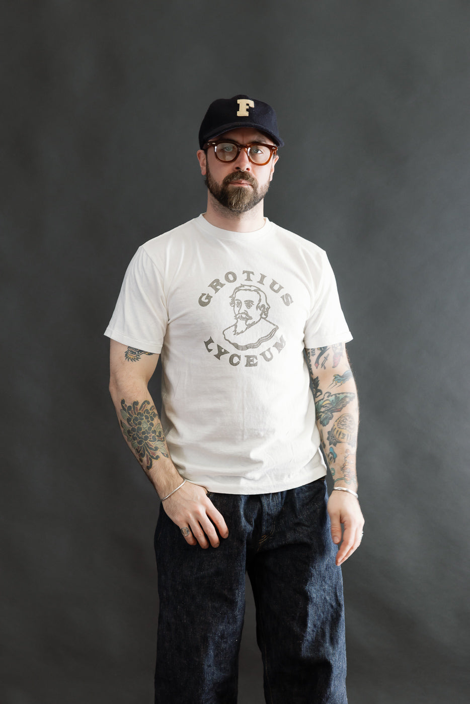 Man wearing the 5.4oz Raffy Tenjiku Printed T-Shirt Grotius - Off White, showcasing the line-drawing portrait design, paired with dark indigo jeans and silver accessories against a neutral backdrop.