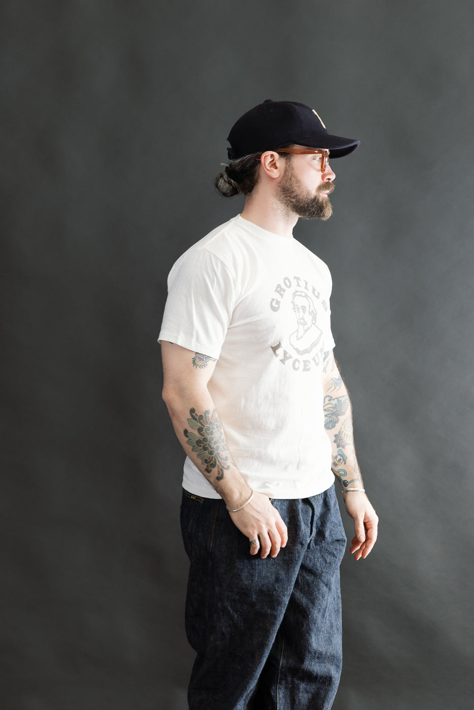 A bearded man models the 5.4oz Raffy Tenjiku Printed T-Shirt Grotius - Off White, showcasing its vintage-inspired fit and tubular construction, paired with loose-fit dark jeans and accessories.