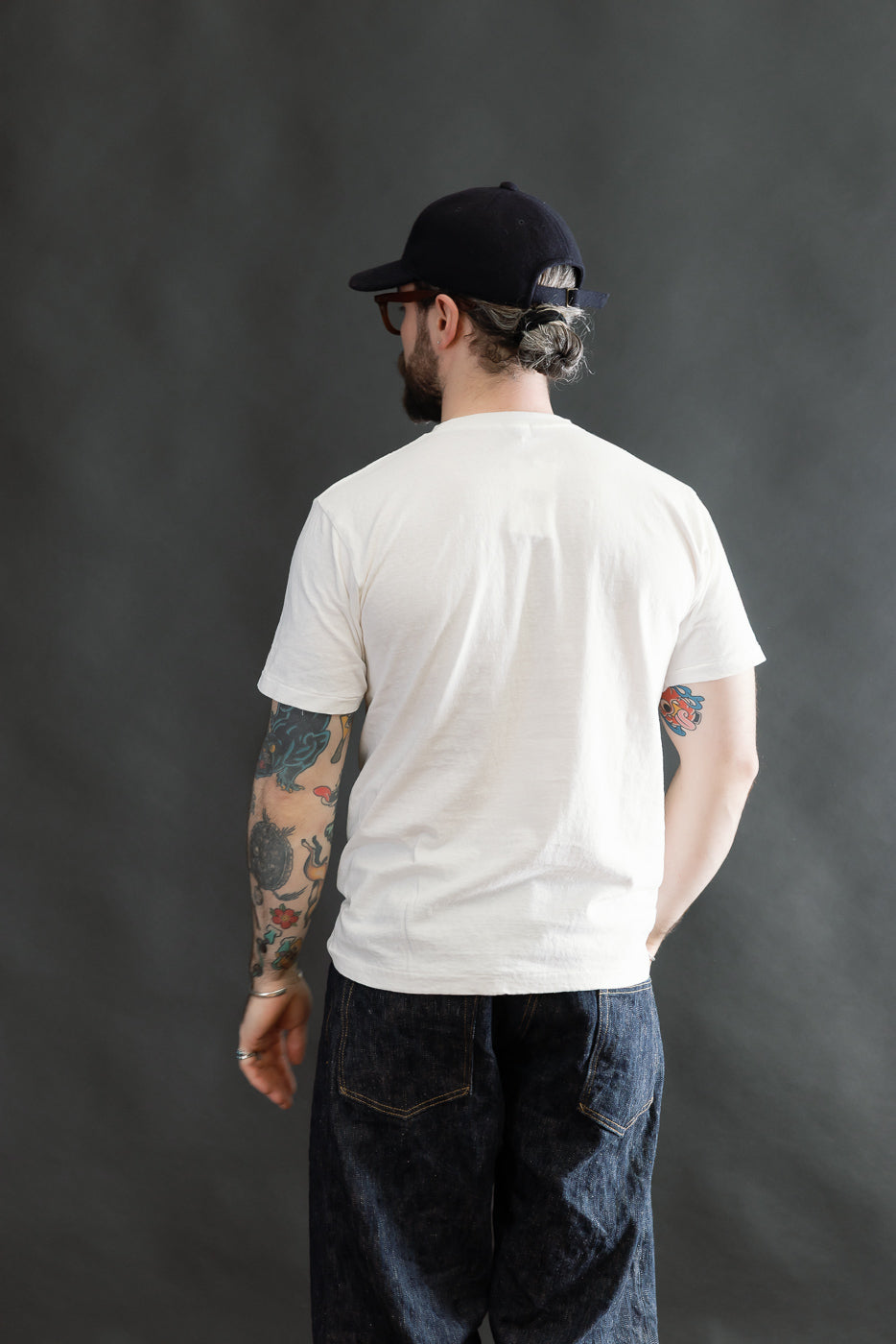 The 5.4oz Raffy Tenjiku Printed T-Shirt Grotius in Off White features a vintage-inspired design, worn by a model showcasing colorful tattooed forearms and paired with dark denim jeans and a backward cap.