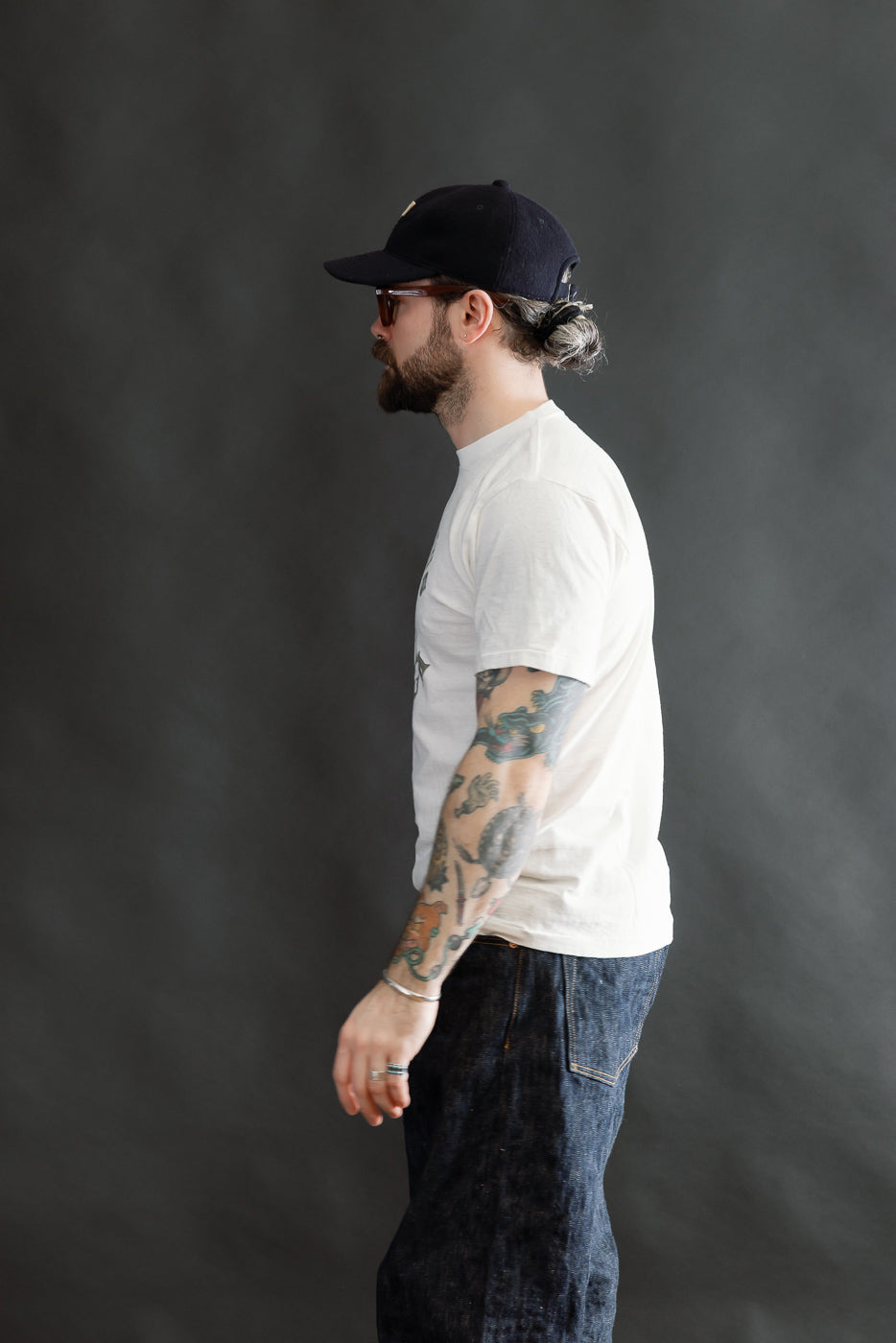 A bearded man in profile wears the 5.4oz Raffy Tenjiku Printed T-Shirt Grotius - Off White, styled with dark denim jeans, showcasing its vintage-inspired, seamless design and textured Japanese cotton fabric.