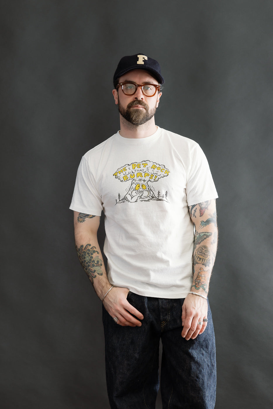 Bearded man wearing a 5.4oz Raffy Tenjiku Printed T-Shirt The Pet Rock - Off White, showcasing colorful tattoos, styled with navy cap, glasses, dark denim jeans, and rings, reflecting Dant's heritage fashion ethos.