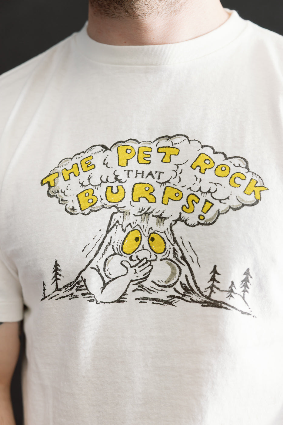 The 5.4oz Raffy Tenjiku Printed T-Shirt The Pet Rock - Off White features a playful cartoon design of a rock creature with yellow eyes and a burp cloud, capturing vintage-inspired style.
