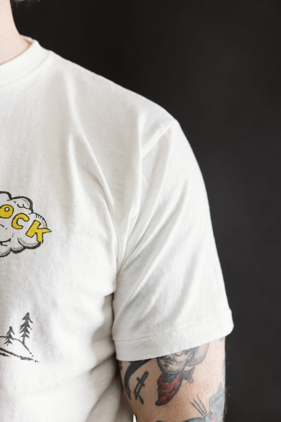 Close-up of a person wearing the 5.4oz Raffy Tenjiku Printed T-Shirt The Pet Rock - Off White, showcasing a cloud graphic with OCK and a tree scene, alongside colorful traditional tattoos.