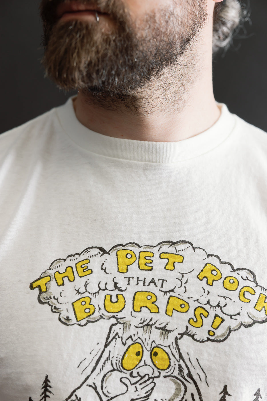 Bearded person wearing the 5.4oz Raffy Tenjiku Printed T-Shirt The Pet Rock in Off White, showcasing a playful cartoon graphic with a burping rock character, emphasizing vintage-inspired design and premium craftsmanship.