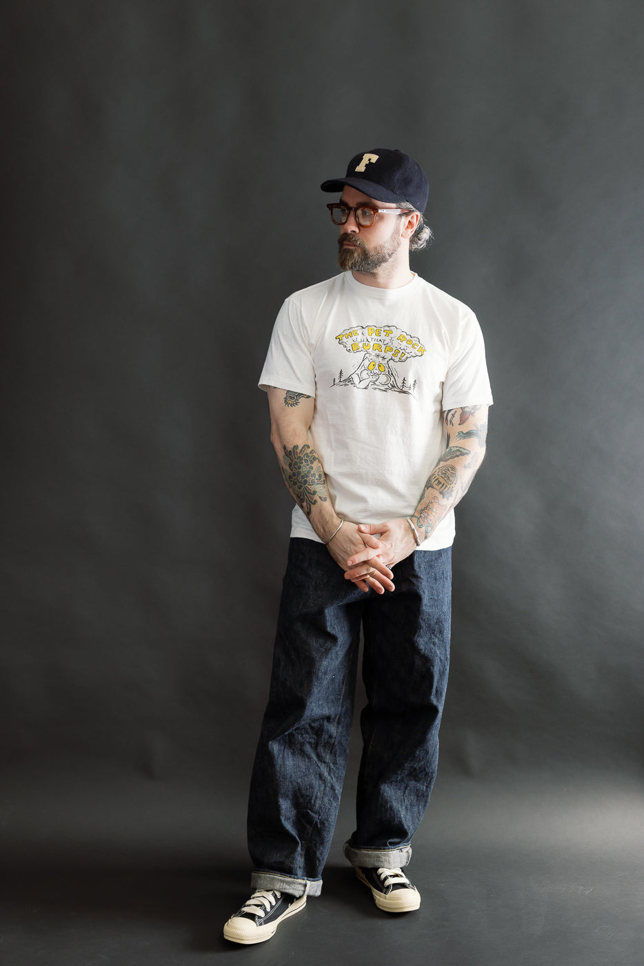 A man models the 5.4oz Raffy Tenjiku Printed T-Shirt The Pet Rock - Off White, featuring a vintage-style cartoon print, loose dark denim jeans, and canvas sneakers, embodying Dant's heritage-inspired fashion.