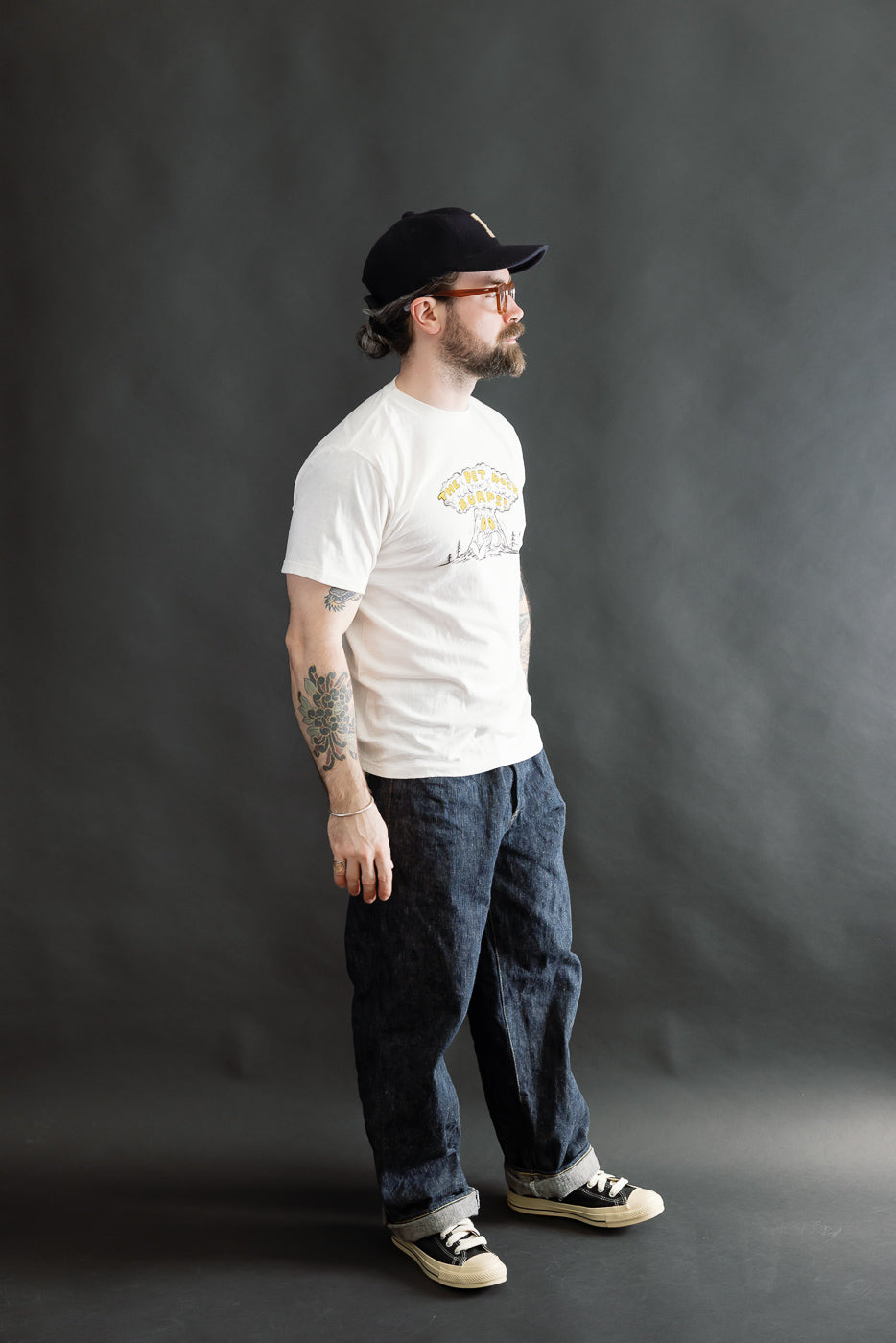 A 5'9 Caucasian male in a 5.4oz Raffy Tenjiku Printed T-Shirt The Pet Rock - Off White, styled with dark blue jeans, black canvas sneakers, and a black baseball cap.