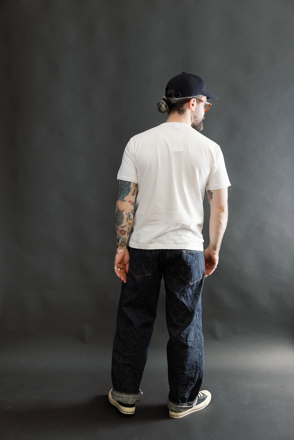 Man in a white 5.4oz Raffy Tenjiku Printed T-Shirt The Pet Rock - Off White, showcasing the tubular knit and tape binder neck, paired with dark jeans and sneakers.