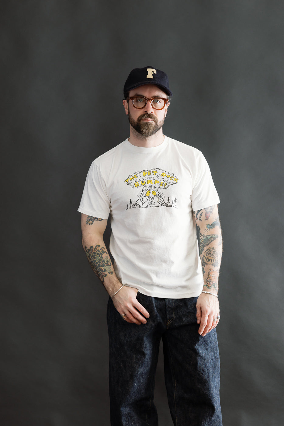 5.4oz Raffy Tenjiku Printed T-Shirt The Pet Rock - Off White, worn by a tattooed man. The T-shirt features a cartoon tree with a speech bubble, paired with dark blue jeans and a black cap.