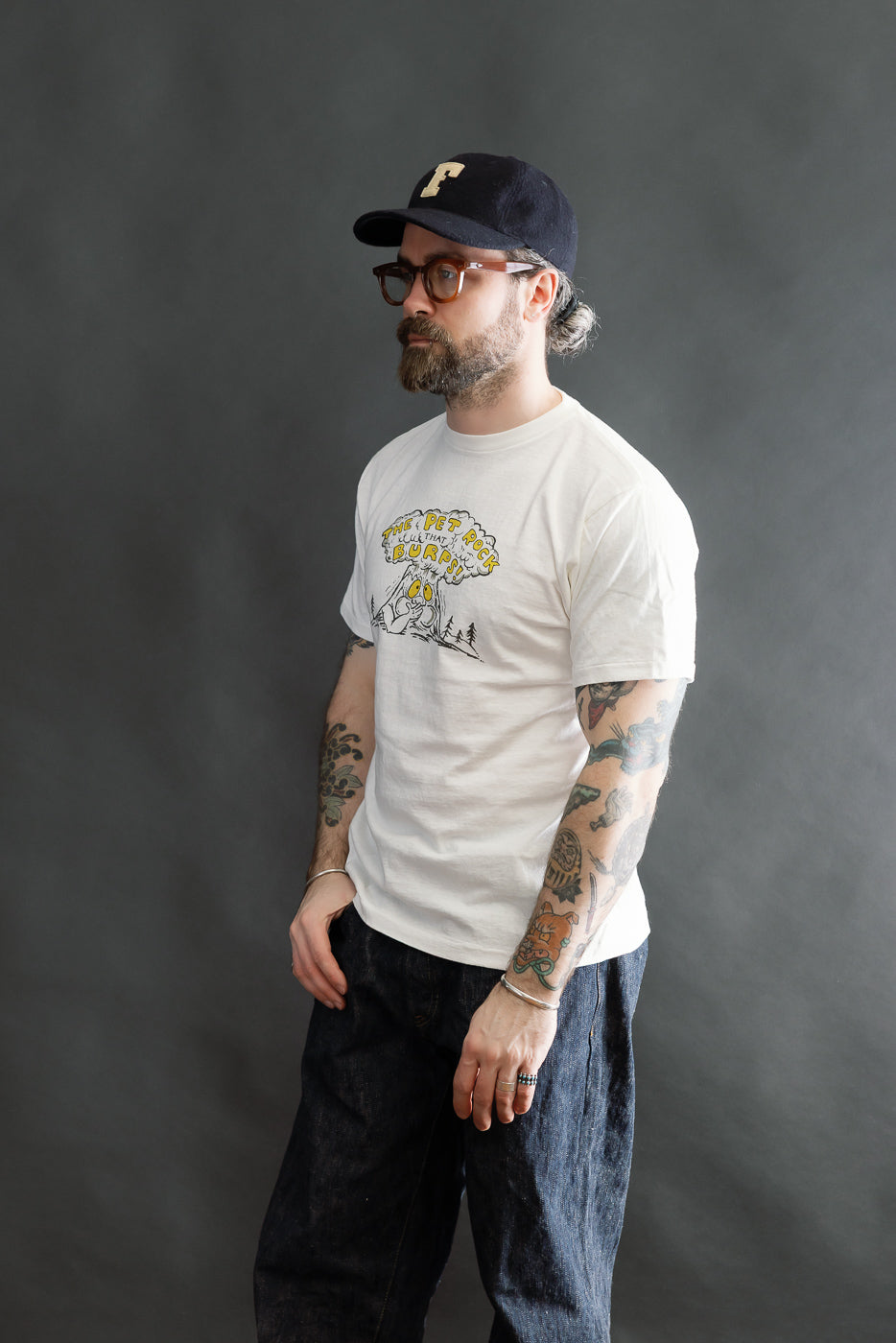 A bearded man models the 5.4oz Raffy Tenjiku Printed T-Shirt The Pet Rock in off-white, showcasing its vintage-inspired design, cartoonish graphic, and tubular knit fit, highlighting heritage craftsmanship.