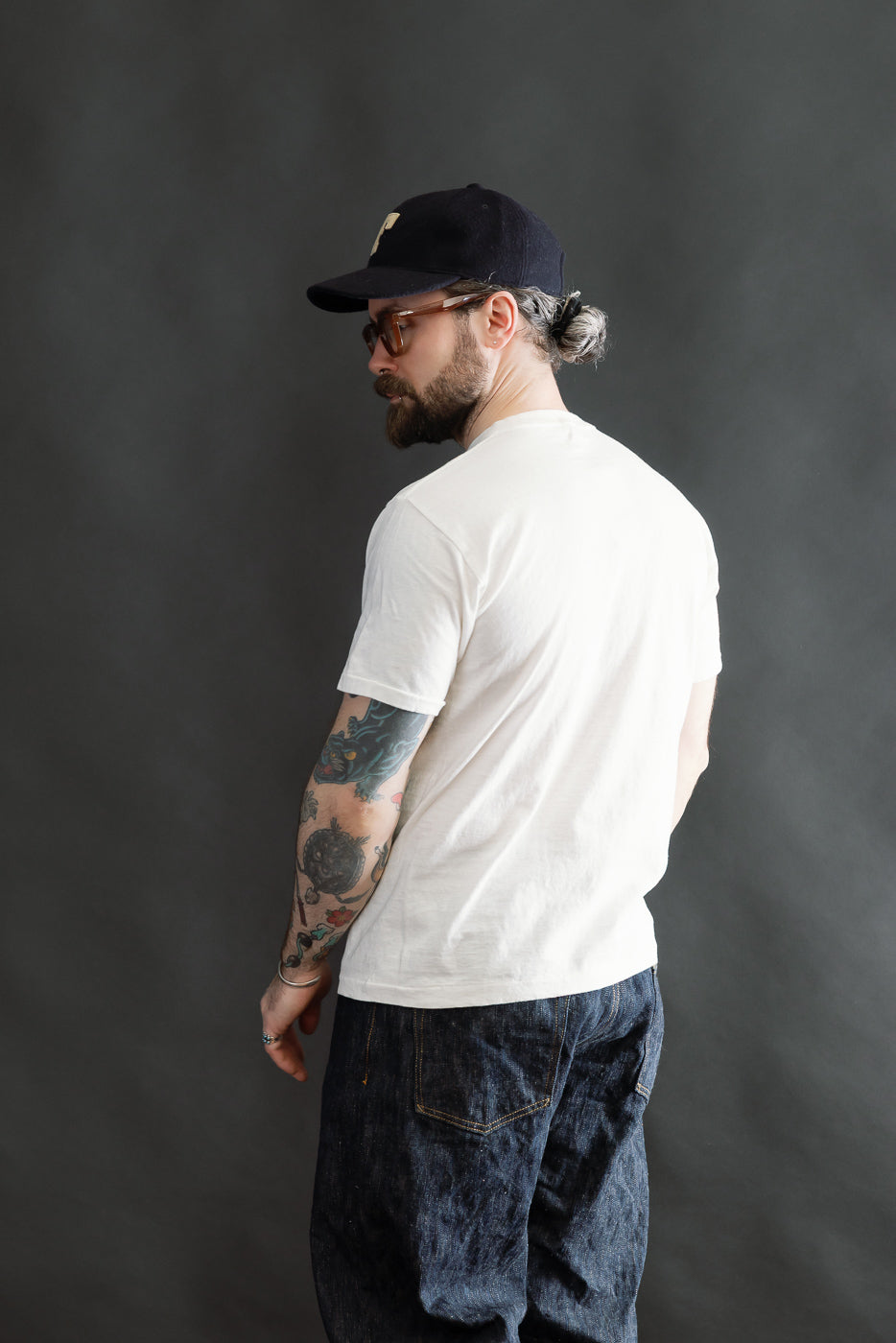 Bearded man in a white 5.4oz Raffy Tenjiku Printed T-Shirt The Pet Rock - Off White, showcasing colorful tattoos on his arm, paired with dark blue jeans and a black cap, embodying vintage American style.