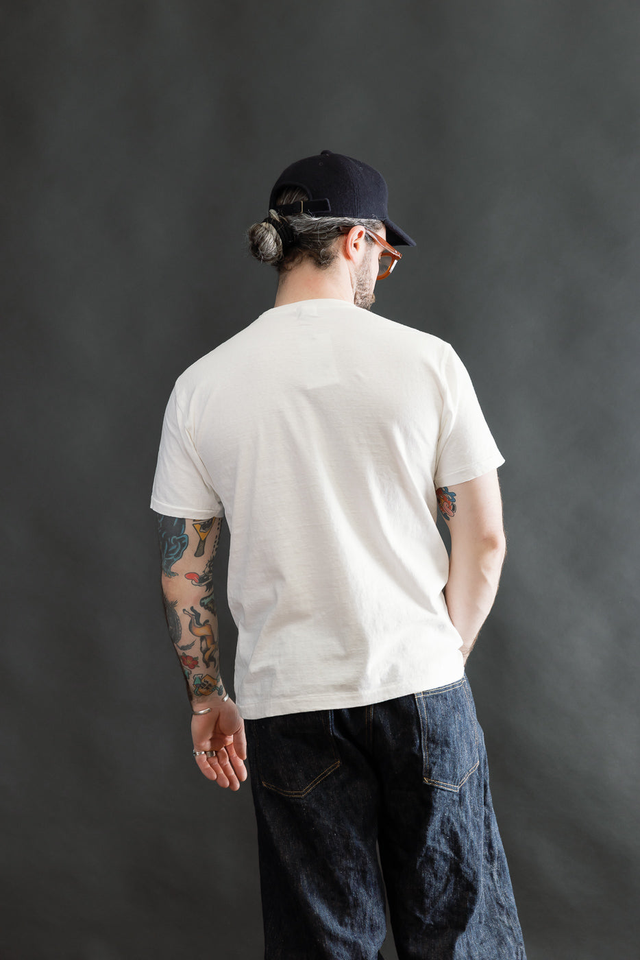 Man wearing the 5.4oz Raffy Tenjiku Printed T-Shirt The Pet Rock in Off White, styled with loose dark denim jeans, showcasing colorful tattooed forearms and accessorized with a black cap and sunglasses.