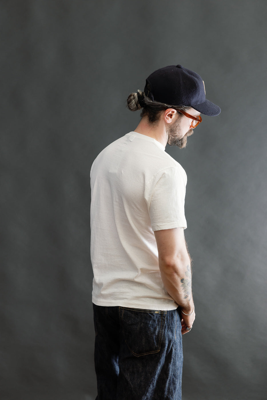 Model wearing 5.4oz Raffy Tenjiku Printed T-Shirt The Pet Rock in off-white, showcasing its vintage-inspired slub jersey texture and seamless tubular construction, styled with dark jeans and a baseball cap.