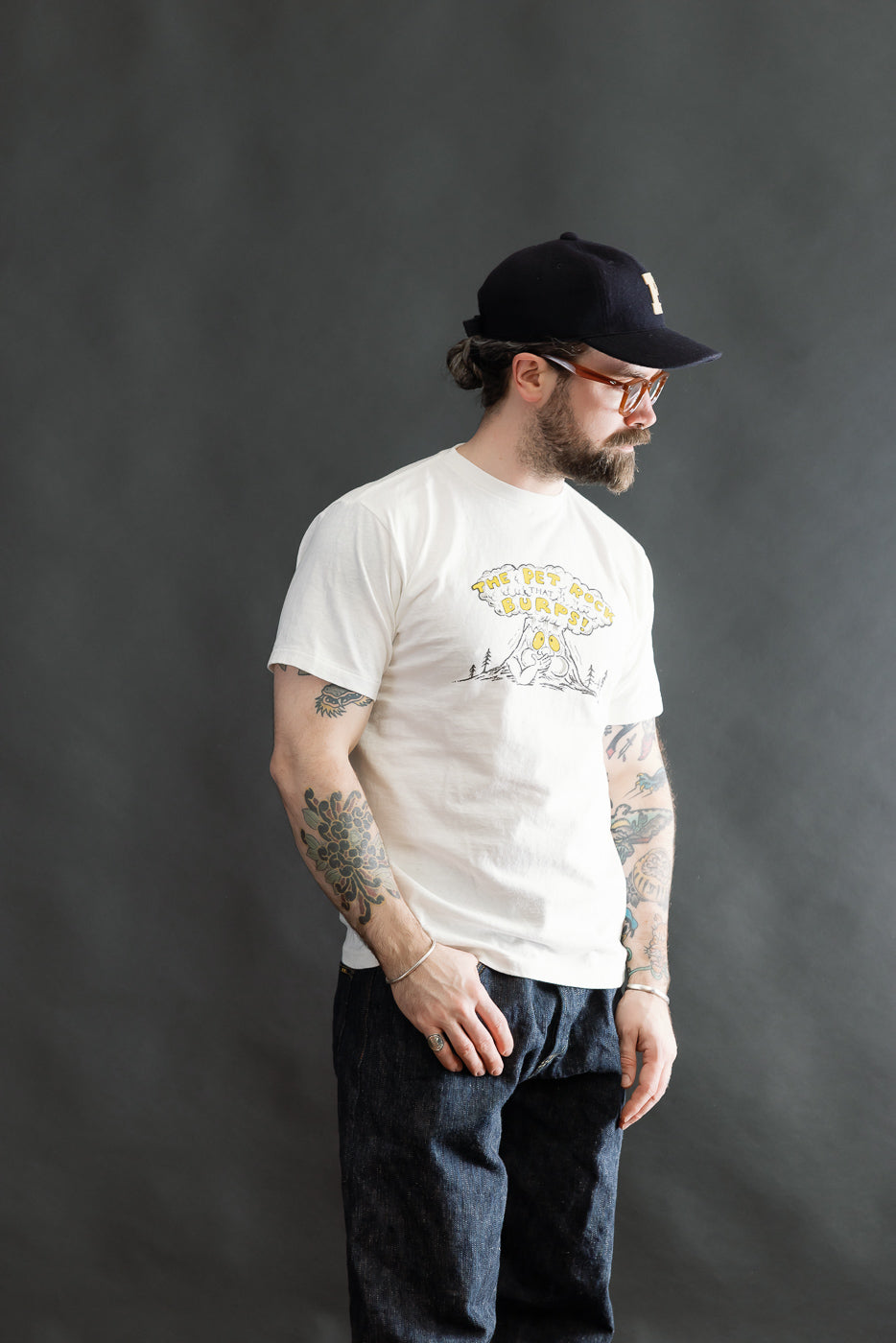 Male model wearing the 5.4oz Raffy Tenjiku Printed T-Shirt The Pet Rock - Off White, styled with dark denim trousers and a black cap, showcasing Dant's heritage-inspired menswear.