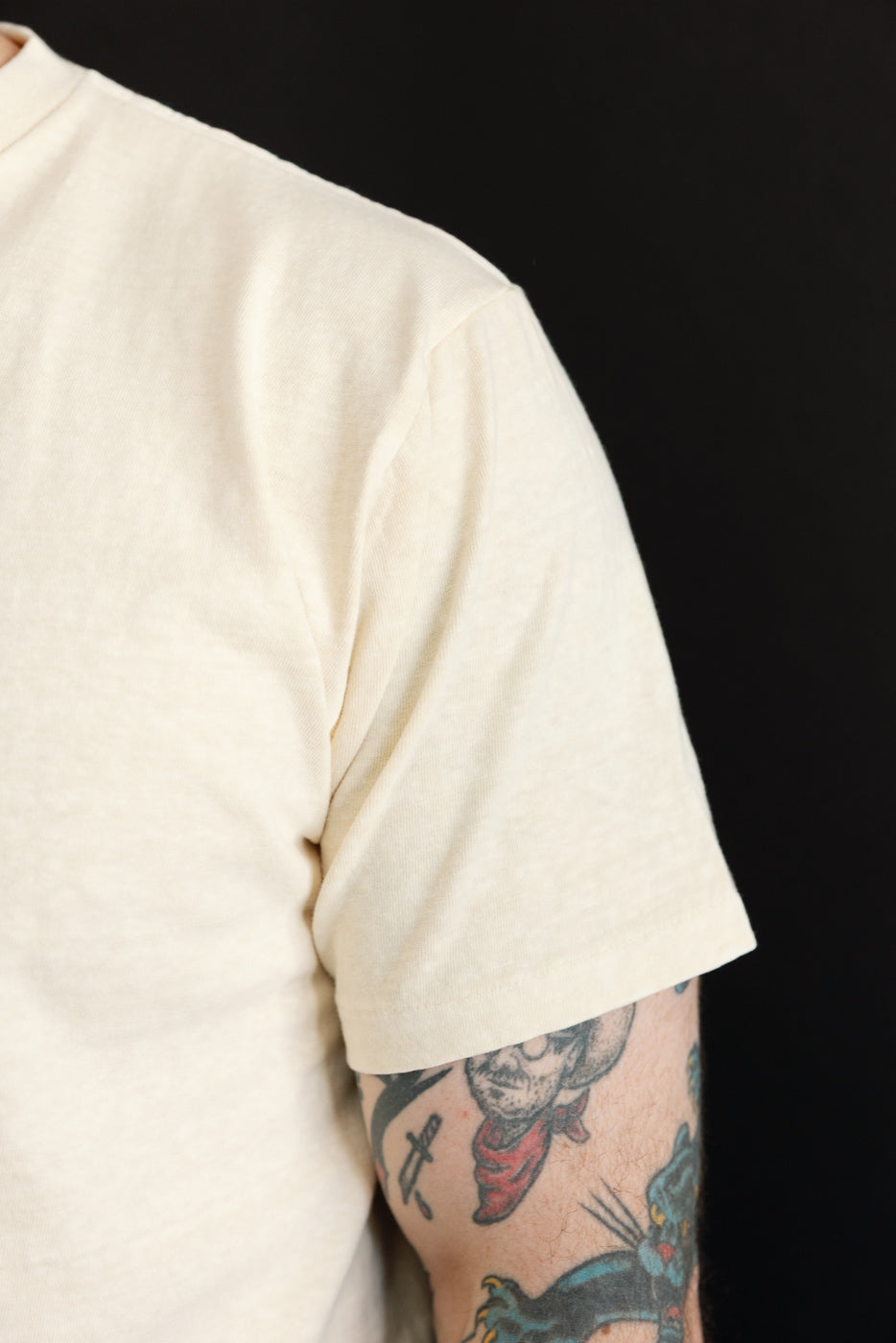 Close-up side view of male model’s shoulder and sleeve, cream tee with ribbed crew neck, athletic build