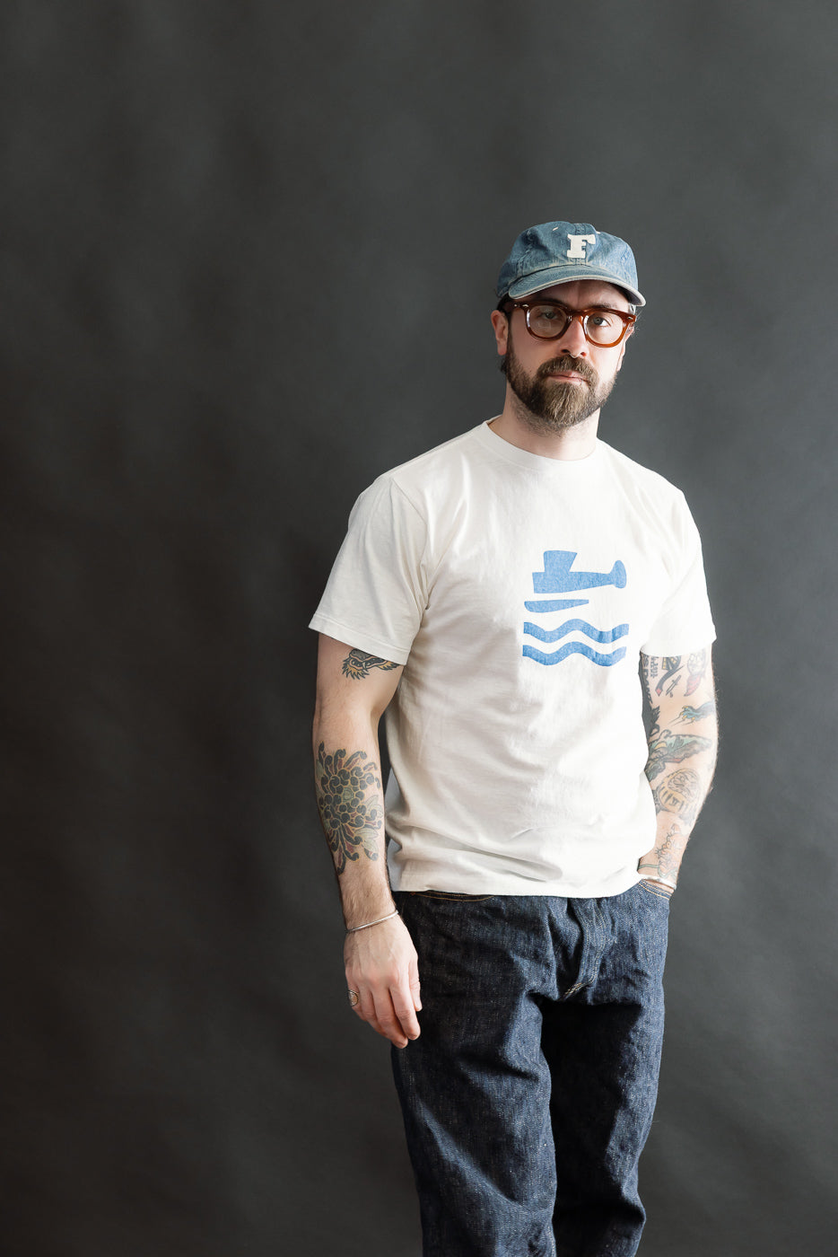 Man wearing 5.4oz Raffy Tenjiku Printed T-Shirt Seaplane - Off White, featuring a blue boat graphic, paired with dark denim jeans, blue baseball cap, and round glasses, against a plain backdrop.