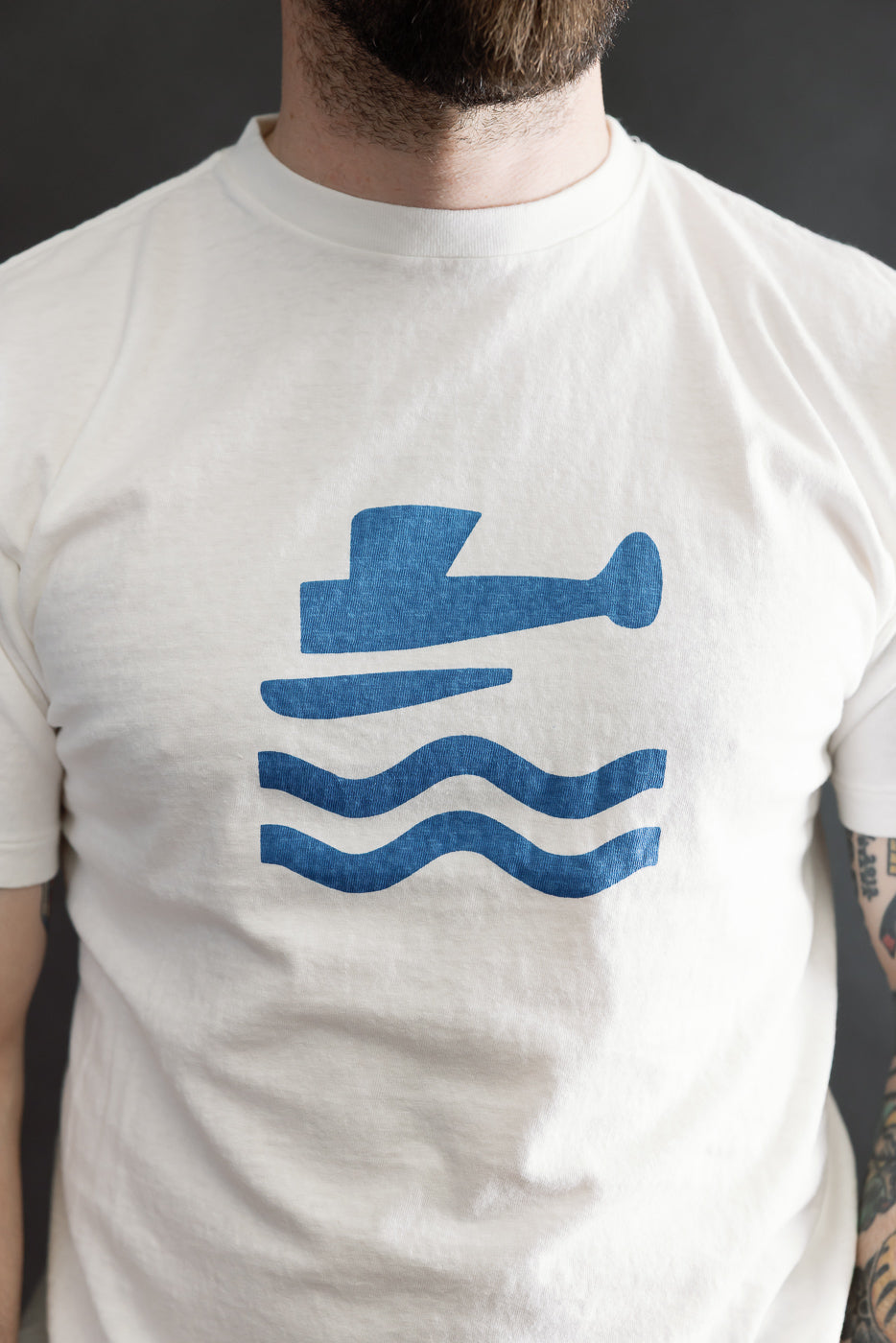 A bearded person wears the 5.4oz Raffy Tenjiku Printed T-Shirt Seaplane - Off White, showcasing its blue floatplane graphic and water waves on a slub jersey fabric. A tattooed arm is partially visible.