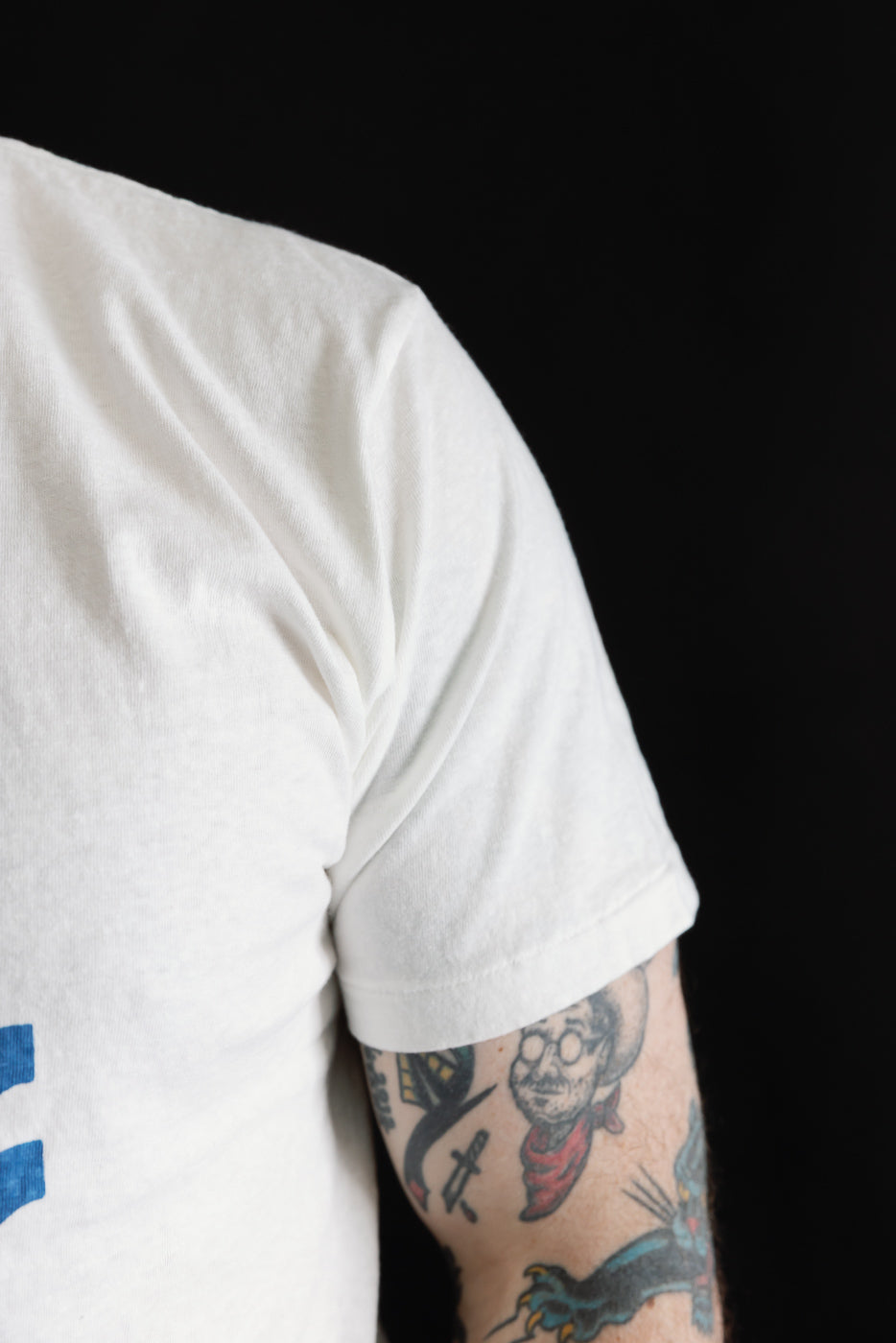 The image features the 5.4oz Raffy Tenjiku Printed T-Shirt Seaplane - Off White, showcasing its seamless tubular knit design. The fabric's nep-like texture is highlighted, contrasting against a tattooed forearm.