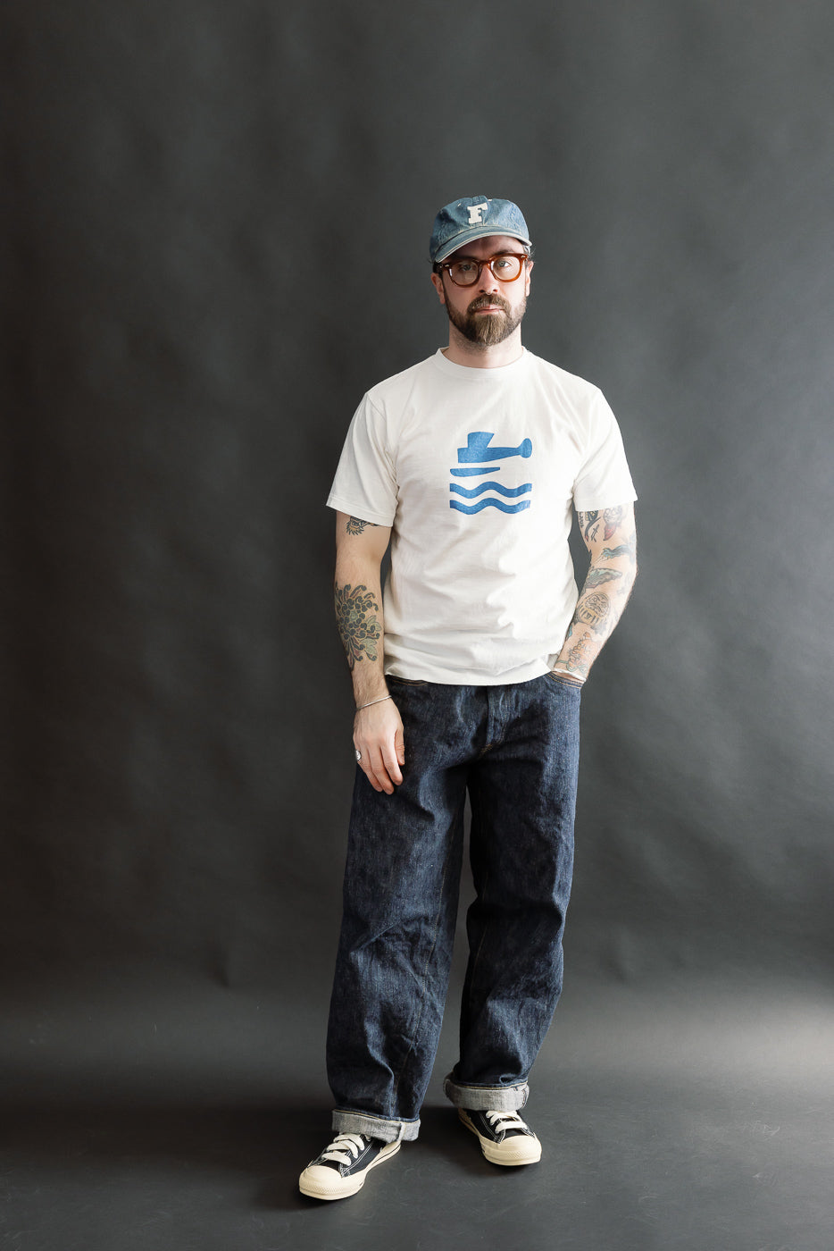 A 5.4oz Raffy Tenjiku Printed T-Shirt Seaplane in Off White is worn by a male model. The T-shirt features a blue graphic and complements dark blue jeans and black-and-white sneakers.