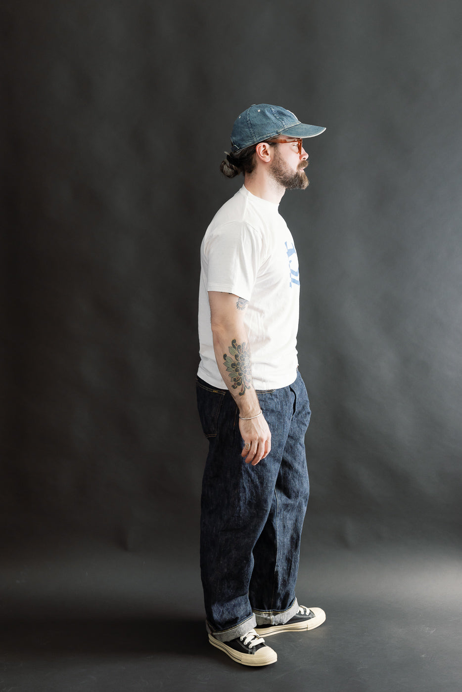 A 5'9 man wears a 5.4oz Raffy Tenjiku Printed T-Shirt Seaplane in off-white, styled with rolled-up dark denim jeans and black-and-white sneakers, showcasing a relaxed and minimalist look.