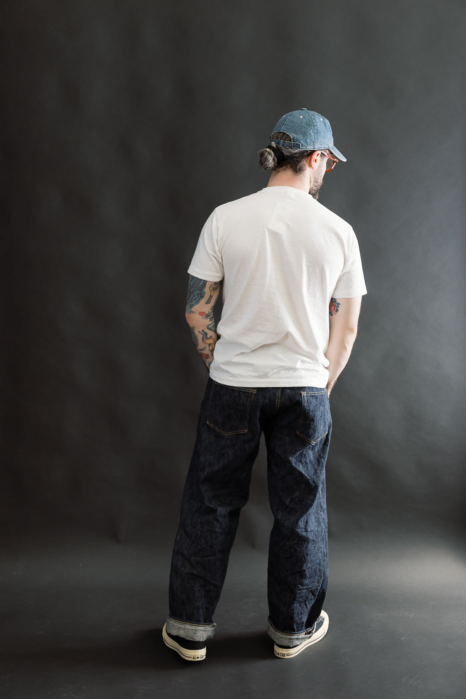Man in a 5.4oz Raffy Tenjiku Printed T-Shirt Seaplane - Off White, dark indigo jeans, low-top sneakers, and a backward blue denim cap, showcasing a heritage-inspired look with visible arm tattoos.